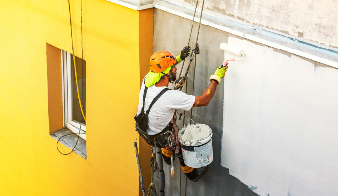 a person working on an exterior wall, applying paint from a bucket using a roller